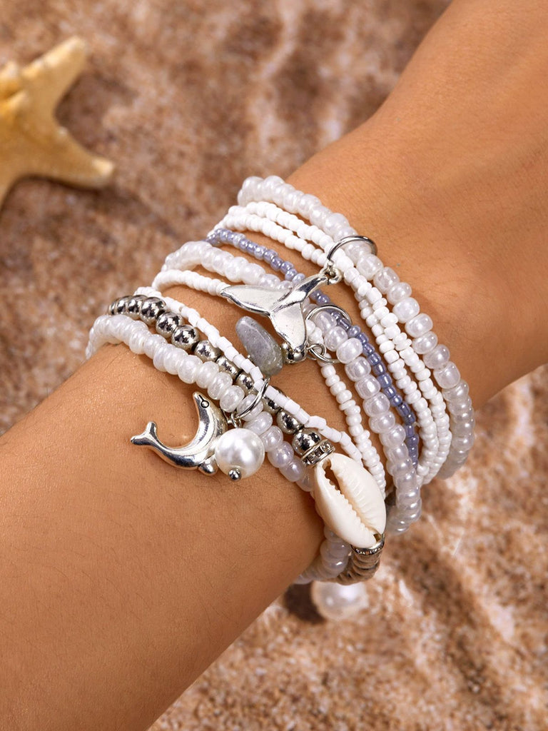 Plusieurs bracelets de perles ornés de coquillages et de breloques en métal, portés au poignet sur un fond sableux.