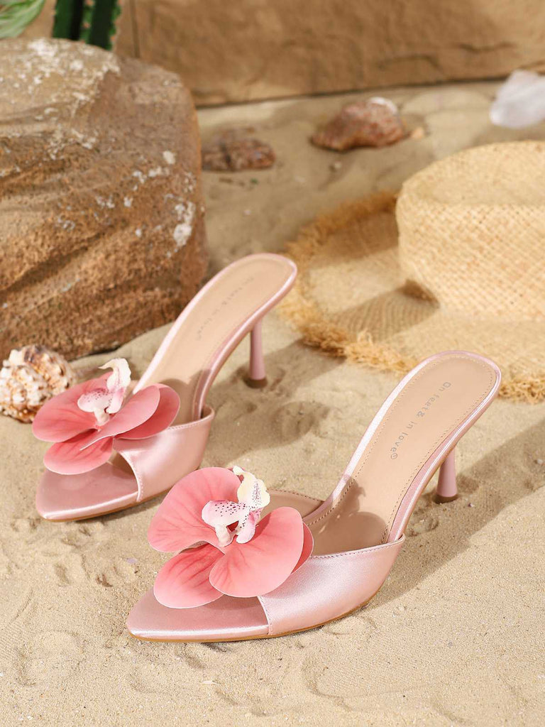 A pair of light pink satin mule heels with matching 3D fabric flowers, pictured on sand next to a straw hat.